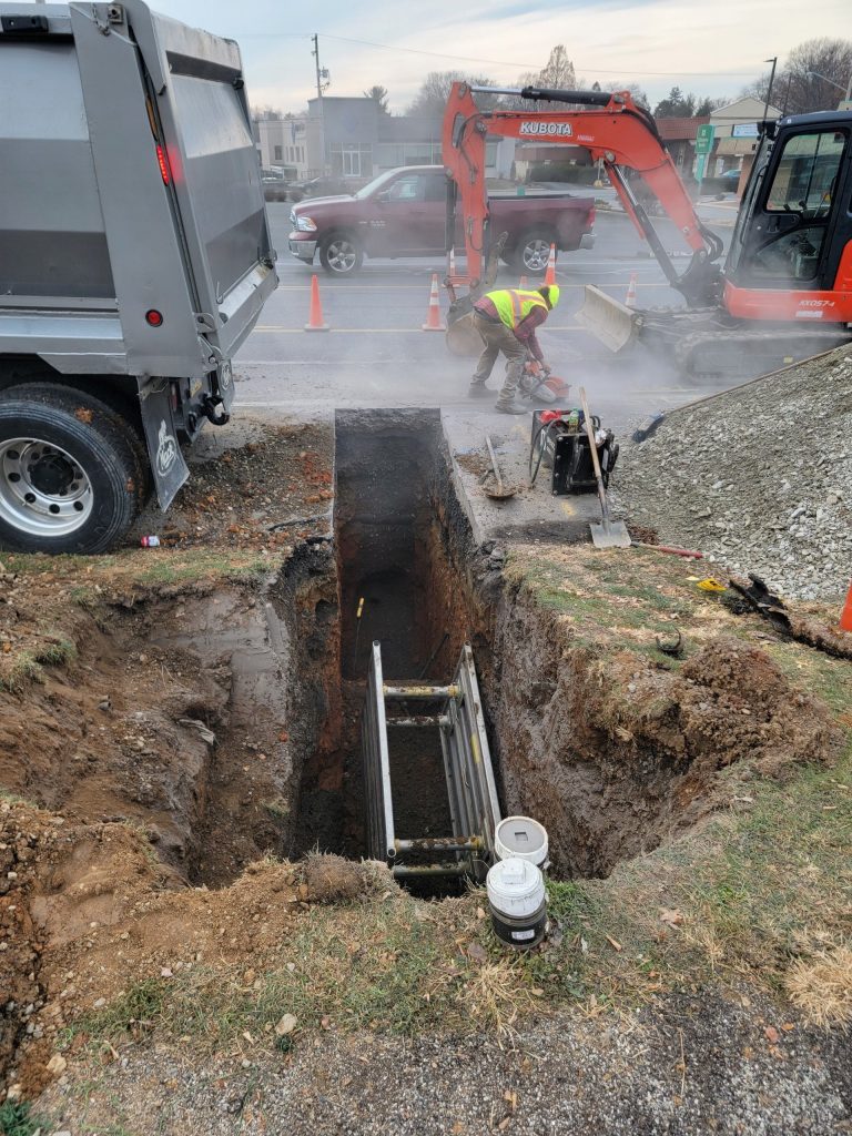 A construction site with a deep trench in the foreground contains a ladder. Workers in safety gear, including someone using power machinery, are on site. An excavator and truck are busy excavating nearby while traffic cones mark the area. Dust or steam fills the air as jetting equipment operates efficiently.