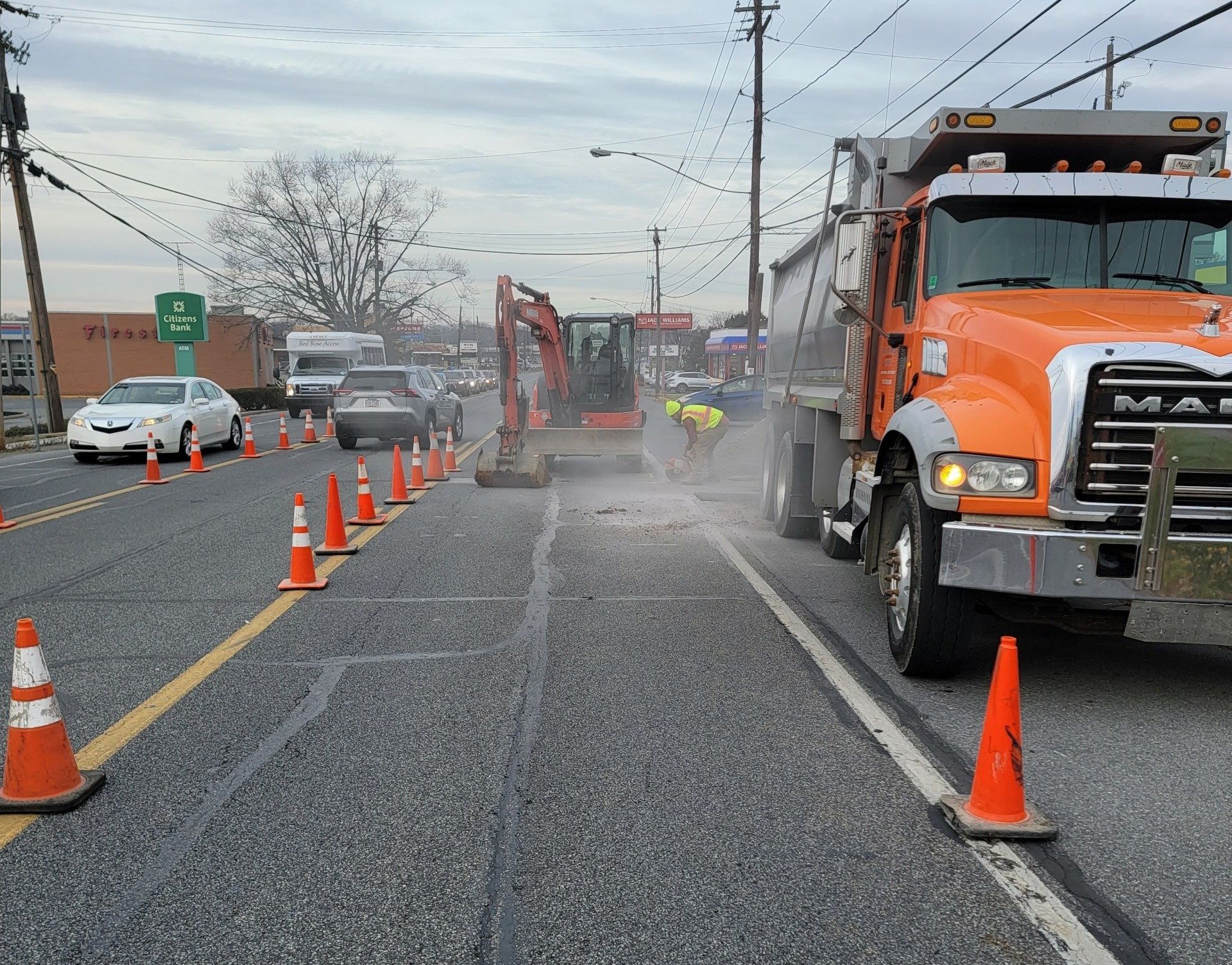 Workers are busy excavating at a road construction scene, operating an excavator on the paved street. Orange cones direct traffic into one lane while a parked orange truck is on the right. Cars queue to the left under an overcast sky, with utility poles in the background.