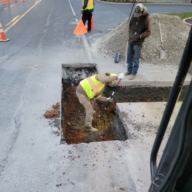 Construction workers are repairing a road. One worker is using a tool in a rectangular hole, while another stands nearby with a shovel. Orange cones ensure safety as gravel piles to the side. Collaborating with Sam's Backhoe & Septic Repairs, they both wear safety vests for protection.