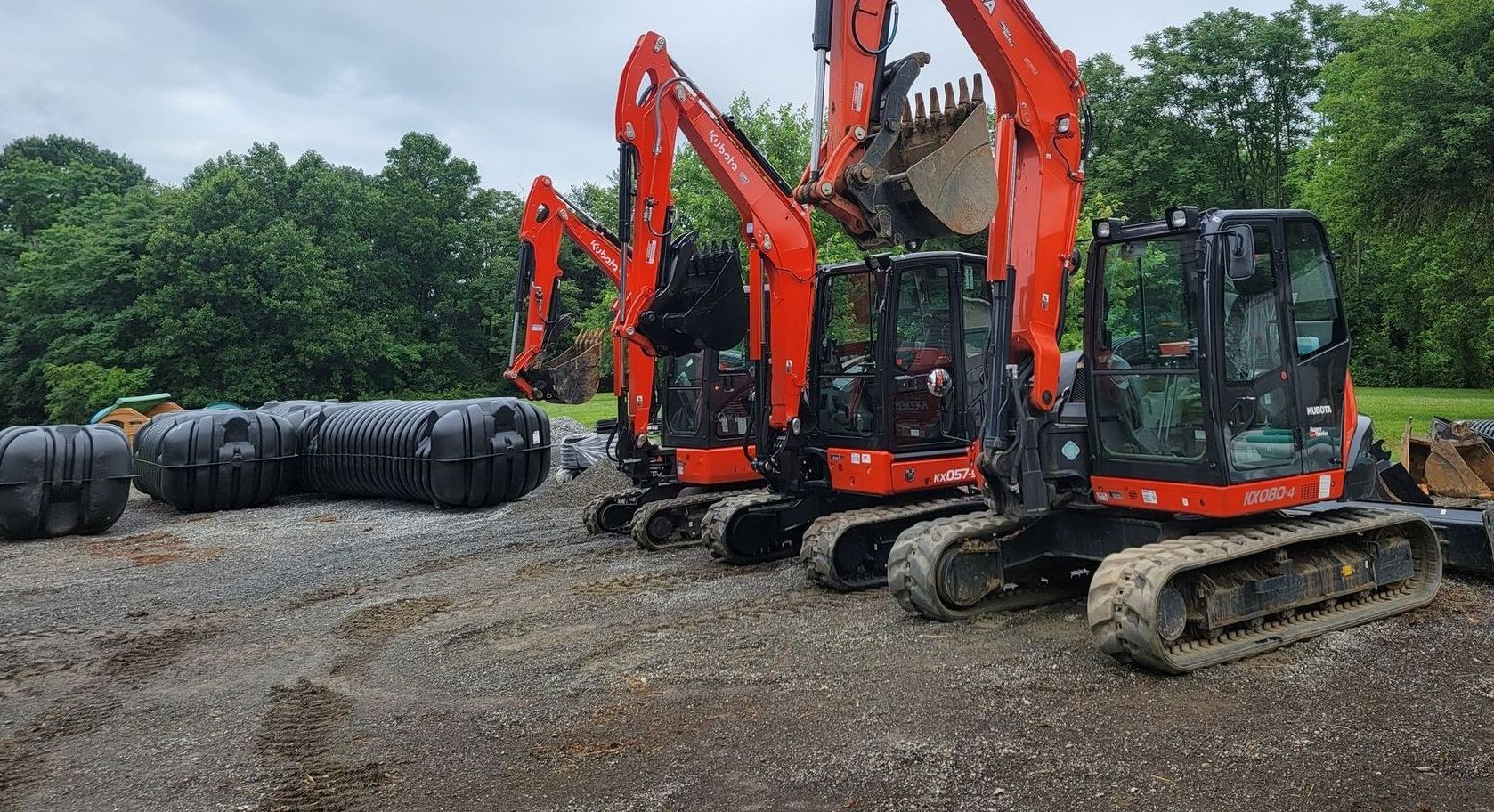 Three orange excavators from Sam's Backhoe & Septic Repairs are parked on a gravel lot next to several large black plastic tanks. Trees and greenery provide a lush backdrop under the cloudy sky, setting the stage for efficient solutions to clogged drains.