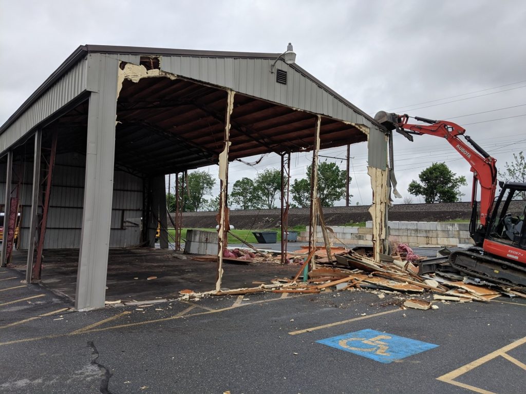 An excavator is demolishing a large metal-framed building with its roof partially intact, while debris is scattered on the ground. In the foreground, a parking space with a wheelchair symbol is visible. The scene resembles the meticulous process of scoping out a new project site.