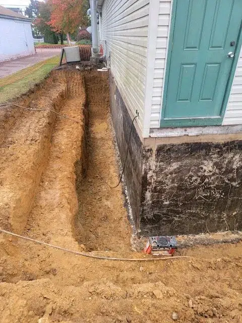 A narrow trench is carefully dug beside a house with white siding and a teal door, running parallel to its foundation. Some construction equipment is visible in the excavating site, and the ground remains bare, potentially indicating work on clogged drains.