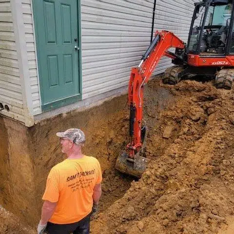 A construction worker from Sam's Backhoe & Septic Repairs stands near a large pile of excavated dirt next to a house with light siding and a green door. An orange excavator is jetting alongside the foundation. The worker is wearing an orange shirt and a camo cap.