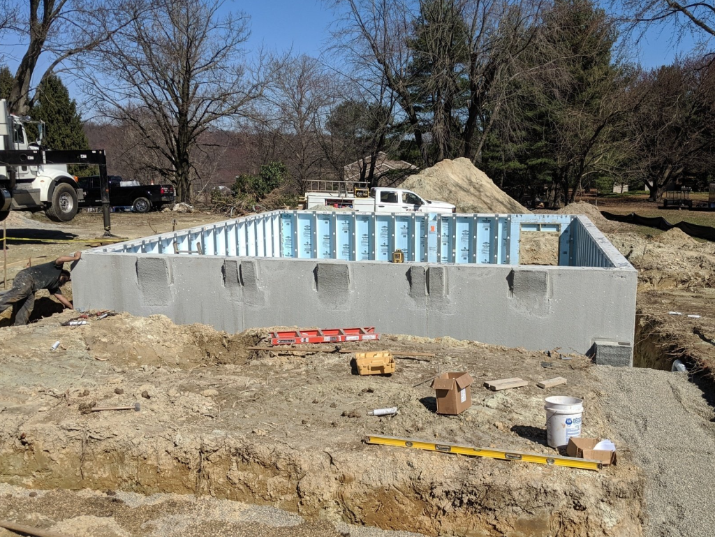 A construction site features a rectangular concrete foundation in progress, surrounded by scattered tools and utility vehicles. An excavating team operates nearby as dirt mounds and materials await use. In the background, trees stand under a clear sky, adding a serene backdrop to the industrious scene.