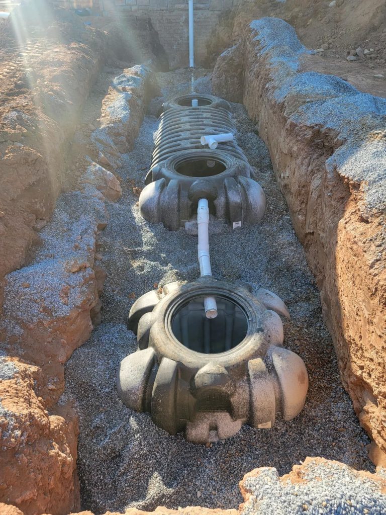 Two large, gray septic tanks are positioned in a freshly dug trench, partially surrounded by gravel. White pipes connect the tanks as sunlight casts rays over the exposed earth of the trench walls.