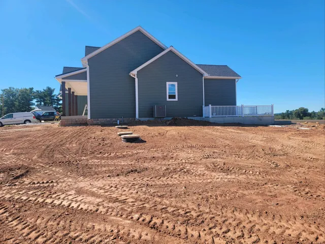 A newly constructed gray house with white trim stands against a clear blue sky. The front features a small porch and a large, freshly graded dirt yard by Sam's Backhoe & Septic Repairs. A white car is parked to the side, and trees are visible in the background.