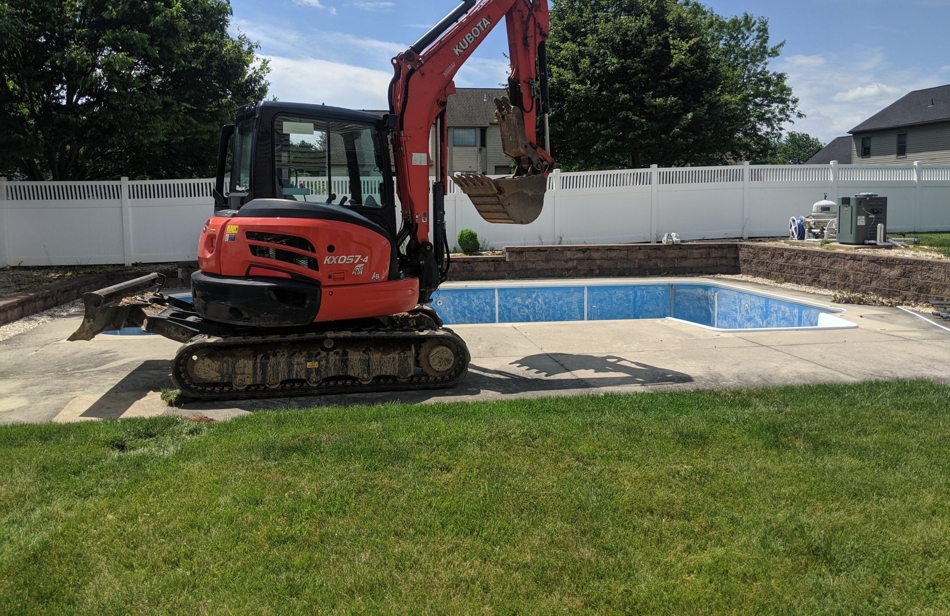 An orange excavator is parked beside an empty in-ground swimming pool in a backyard. The pool, possibly undergoing maintenance for clogged drains, is surrounded by a concrete patio. Lush green trees and a white fence decorate the background under a partly cloudy sky.