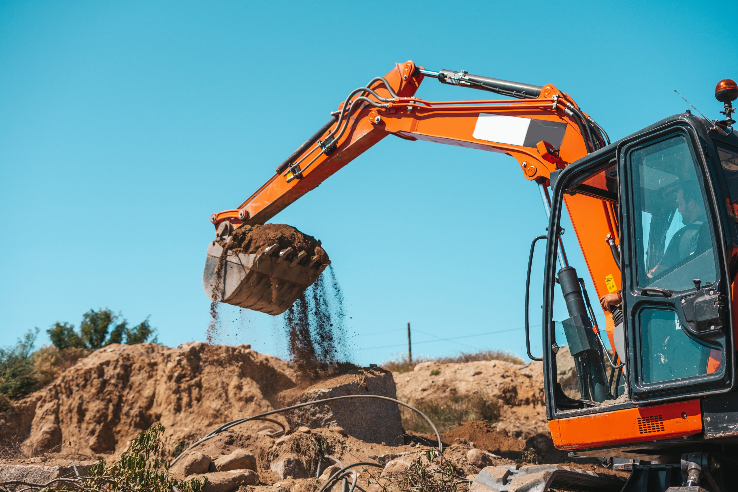 An orange excavator from Sam's Backhoe & Septic Repairs is scooping up dirt and rocks, debris cascading from its bucket. Set against a clear blue sky, it diligently works on a construction site surrounded by piles of earth and sparse vegetation.