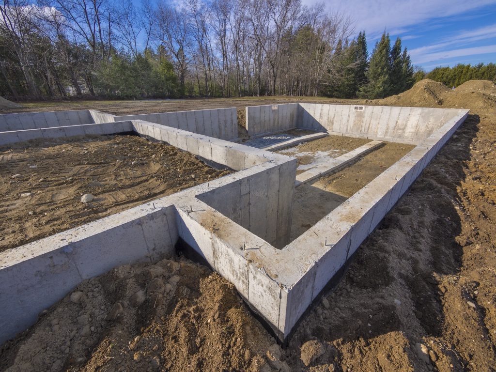 The concrete foundation of a building under construction is surrounded by piles of dirt and bare trees. The structure outlines a rectangular shape, set against a clear blue sky. Careful scoping ensures that future development will avoid issues like clogged drains.