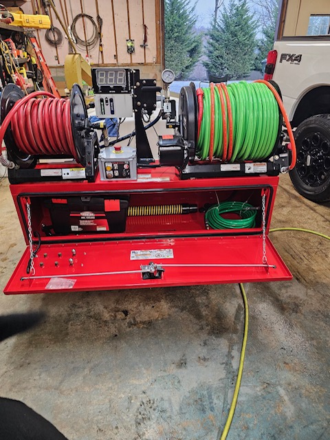 A red machine with two large hose reels, one red and one green, is set up in a garage. It's equipped with gauges, a digital display, and multiple control buttons for scoping purposes. A yellow hose is connected at the bottom. A white vehicle is visible in the background.