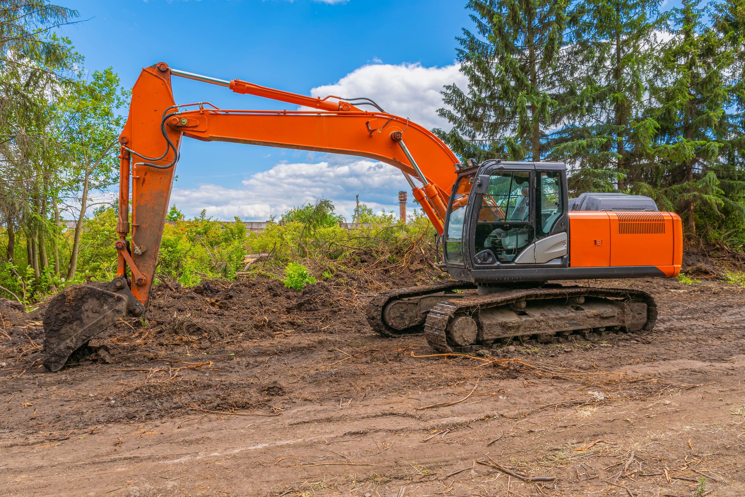 At Sam's Backhoe & Septic Repairs, an orange excavator rests on a dirt path at a construction site. It's surrounded by trees and piles of soil under a partly cloudy blue sky, ready for any excavating tasks.