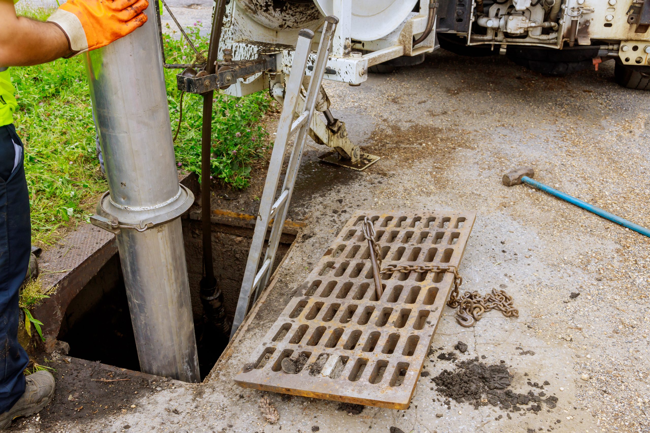 A worker operates machinery to clean clogged drains in a sewer. A metal grate rests on the ground beside a hole with a large pipe inserted, while a ladder leans against the equipment. A hammer is visible on the asphalt, with grass and parts of a truck seen in the background.