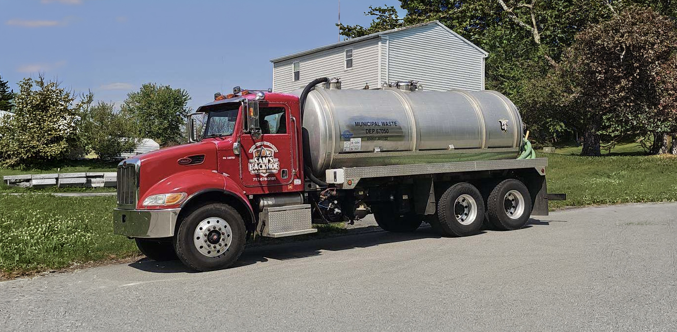 A red truck with a large silver tank labeled “Municipal Waste” from Sam's Backhoe & Septic Repairs is parked on a residential street near grass and trees, with a white house in the background under a clear sky.