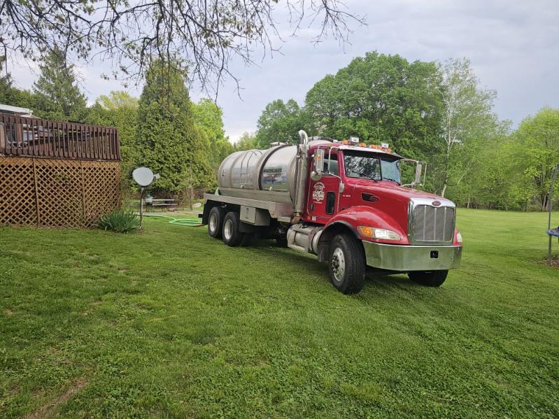 A red tanker truck from Sam's Backhoe & Septic Repairs is parked on a grassy lawn near a wooden deck and satellite dish, with green trees and a cloudy sky in the background.