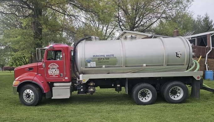 A red and silver municipal waste septic truck from Sam's Backhoe & Septic Repairs is parked on grass near a house, with green hoses attached at the back and trees in the background.