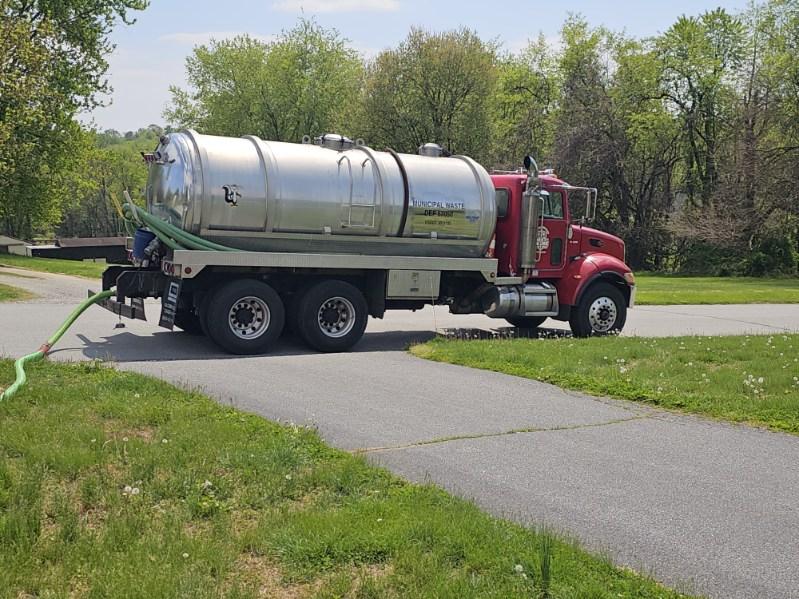 A red and silver Sam's Backhoe & Septic Repairs service truck is parked on a residential street with a green hose extending from the tank. Green grass and trees surround the scene on this sunny day.