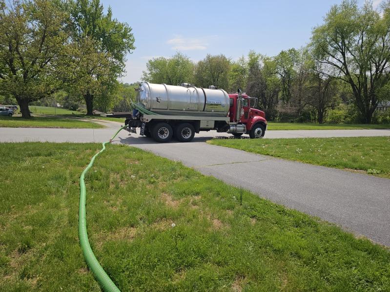 A red and silver tanker truck from Sam's Backhoe & Septic Repairs is parked on a paved road beside a grassy area, with a long green hose extending from the truck across the grass under a sunny sky.