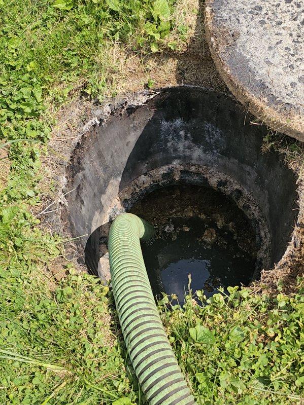 A green-striped hose inserted into an open, round concrete septic tank surrounded by grass, with the tank’s lid partially removed and resting on the grass, showcases a typical service visit from Sam's Backhoe & Septic Repairs.